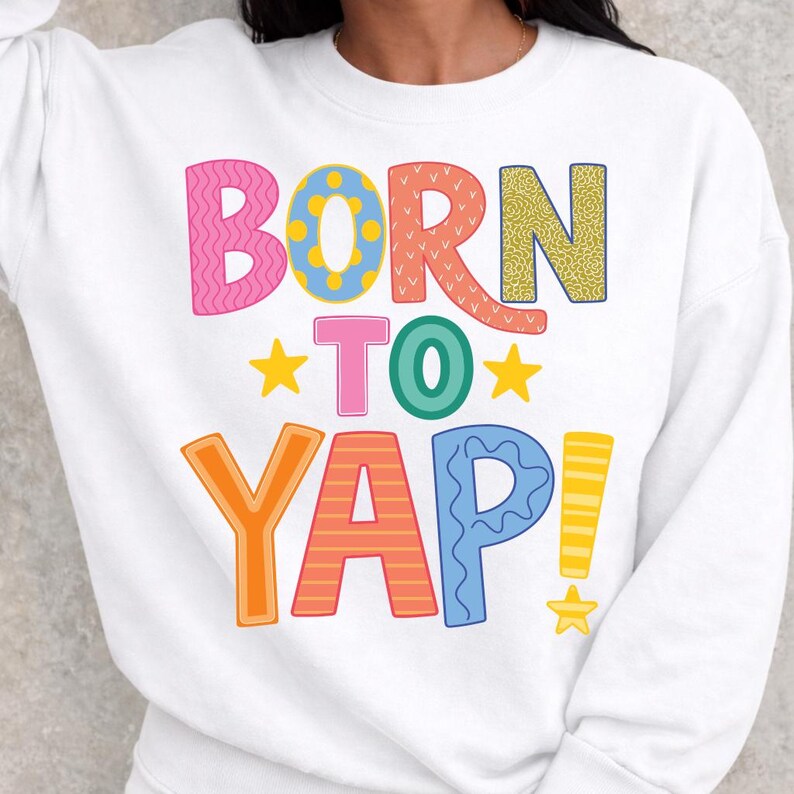 May include: White sweatshirt with the phrase "BORN TO YAP" in colorful, playful lettering. The text features various patterns and colors, including pink, blue, orange, and yellow, with star accents.