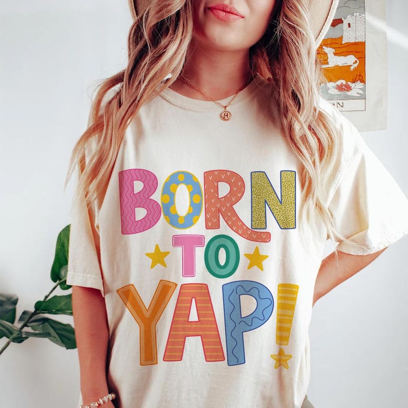 May include: Cream-colored t-shirt with the phrase "BORN TO YAP!" in colorful, patterned letters. The design includes stars and exclamation points. The shirt is a casual, everyday wear item.