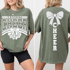 May include: Olive green t-shirt with the word "MASCOT CHEER" in white block letters and a bow graphic. The back of the shirt features a large bow and the word "CHEER" vertically. The shirt is a comfortable, oversized fit.