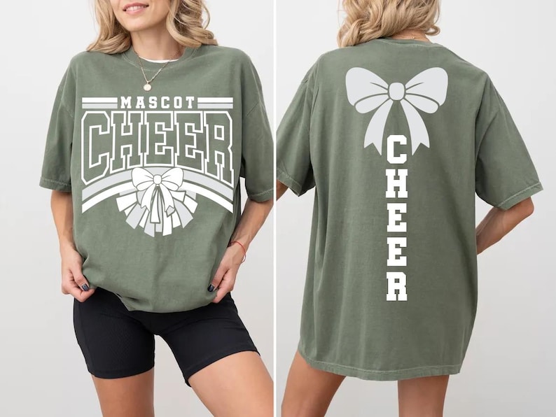 May include: Olive green t-shirt with the word "MASCOT CHEER" in white block letters and a bow graphic. The back of the shirt features a large bow and the word "CHEER" vertically. The shirt is a comfortable, oversized fit.
