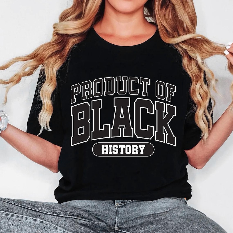 May include: Black t-shirt with the text "PRODUCT OF BLACK HISTORY" in white, outlined letters. The shirt is a crew neck style and is being worn by a person. The person is wearing blue jeans.