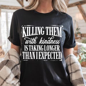 Puede incluir: Una camiseta negra de manga corta con texto blanco que dice: KILLING THEM with kindness IS TAKING LONGER THAN I EXPECTED. Las letras est&aacute;n dispuestas en un bloque rectangular con un efecto de borde desgastado y envejecido en la parte frontal.