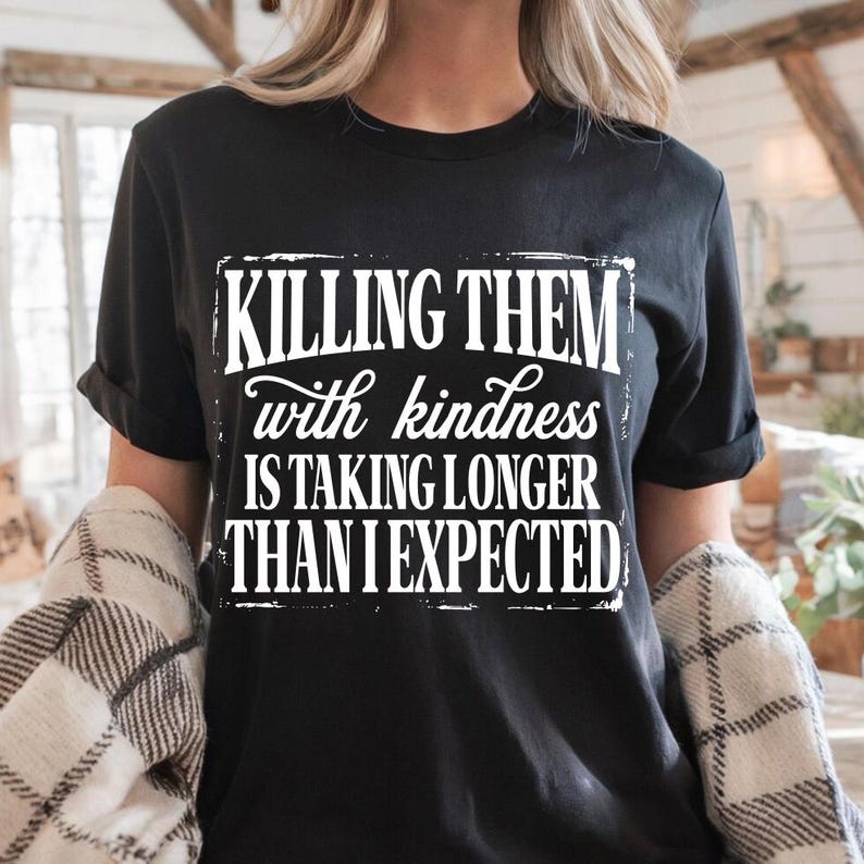Puede incluir: Una camiseta negra de manga corta con texto blanco que dice: KILLING THEM with kindness IS TAKING LONGER THAN I EXPECTED. Las letras est&aacute;n dispuestas en un bloque rectangular con un efecto de borde desgastado y envejecido en la parte frontal.