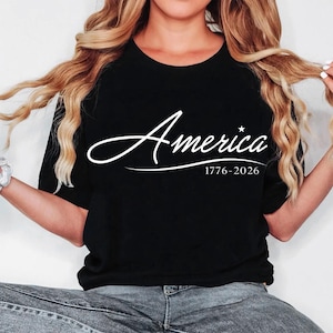 May include: Black t-shirt with the word "America" in a white script font, a star, and the dates "1776-2026" below. The shirt is a crew neck style.