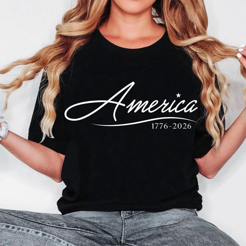 May include: Black t-shirt with the word "America" in a white script font, a star, and the dates "1776-2026" below. The shirt is a crew neck style.
