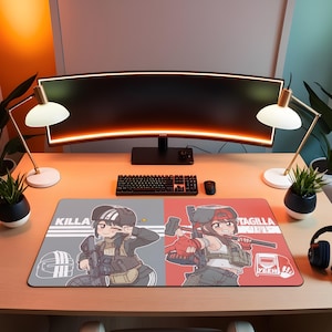 May include: A rectangular gaming mousepad featuring two anime-style characters. One side is gray with a character and the word "Killa", the other is red with a character and the word "Tagilla". The mousepad is on a wooden desk with a keyboard, monitor, and lamps.