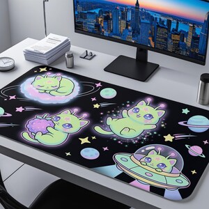 May include: Black desk mat featuring cartoon alien cats in space. The design includes cats in a cloud, holding a berry, floating, and in a UFO, with stars, planets, and a galaxy theme. The mat is rectangular and sits on a white desk.