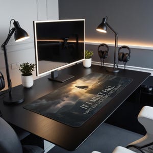 May include: A black desk with a computer monitor, two black desk lamps, and a mouse mat. The mouse mat features a landscape image with the text "IF I MUST FALL I WILL RISE EACH TIME A BETTER MAN."