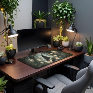 May include: A dark brown desk with a computer monitor, desk lamp, and a mousepad featuring a forest scene with a figure and glowing butterflies. The desk is surrounded by potted plants and a gray office chair.