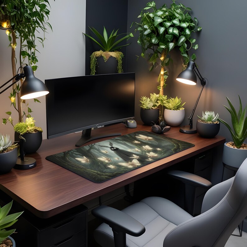 May include: A dark brown desk with a computer monitor, desk lamp, and a mousepad featuring a forest scene with a figure and glowing butterflies. The desk is surrounded by potted plants and a gray office chair.