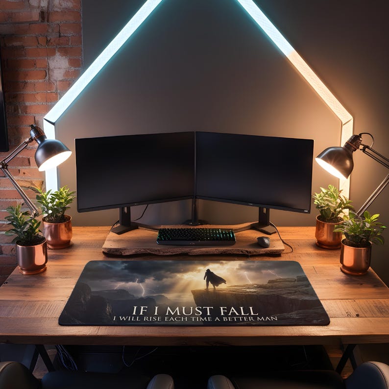 May include: A wooden desk with a mousepad displaying the text "If I Must Fall, I Will Rise Each Time A Better Man" and a dramatic landscape. The setup includes two computer monitors, a keyboard, and two desk lamps.