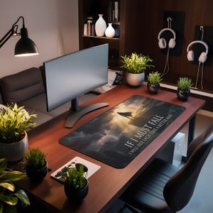 May include: A home office desk with a computer monitor, mouse, and a large rectangular mousepad. The mousepad features a landscape design with a figure and the text "IF I MUST FALL I WILL RISE EACH TIME A BETTER MAN". Several potted plants and headphones are also visible.