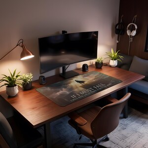 May include: A home office setup with a large curved monitor, speakers, and a desk mat with the text "If I must fall, I will rise each time a better man." The desk is made of dark wood and has a brown leather chair.