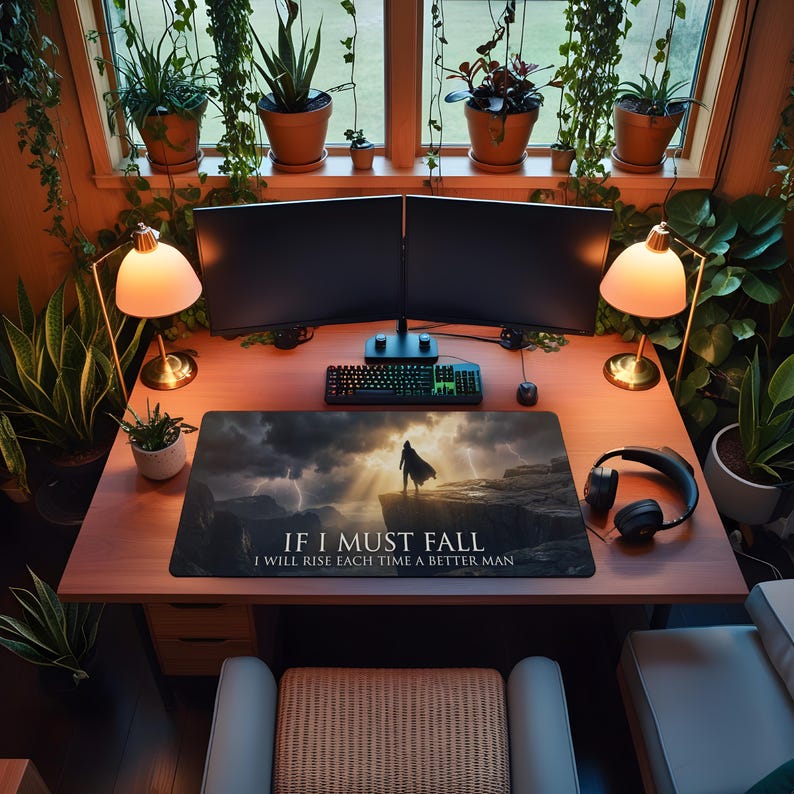 May include: A home office setup featuring a wooden desk with two computer monitors, a keyboard, and a mousepad. The mousepad has the text "If I Must Fall, I Will Rise Each Time A Better Man". Two lamps and headphones are also visible. The room is filled with plants.