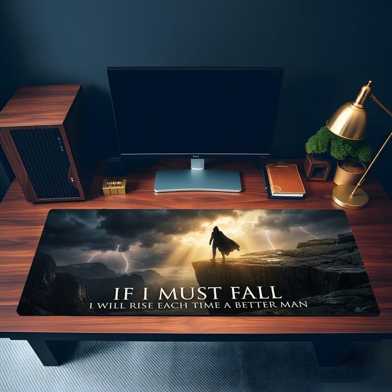 May include: A black rectangular desk mat with a dramatic landscape featuring a cloaked figure on a cliff. The mat displays the text "IF I MUST FALL I WILL RISE EACH TIME A BETTER MAN". The background shows a stormy sky with lightning.