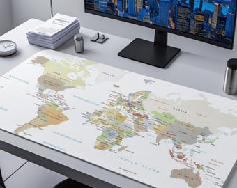 May include: A rectangular desk mat with a world map design, showing continents and oceans in different colours. The mat is on a white desk, with a computer monitor, clock, and other office items in the background. The mat is approximately 80 cm long.