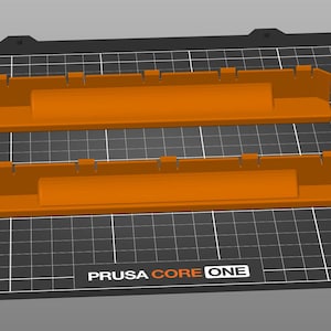 May include: Two orange rectangular 3D-printed objects with notched edges are displayed on a black grid surface. The objects are parallel to each other. The text "PRUSA CORE ONE" is visible on the surface.
