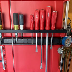 May include: A selection of tools, including screwdrivers with red handles and other implements, arranged on a black rack. The tools are set against a red backdrop, possibly in a workshop or storage area.