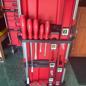 May include: Red tool organiser with a selection of hand tools, including screwdrivers and spirit levels. The tools are arranged in a black holder, with the brand name "Milwaukee" visible on the levels. The organiser is red and has a robust design.