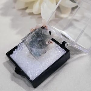 K&ouml;nnte beinhalten: Ein blau-graues Mineralexemplar mit rosa Akzenten, pr&auml;sentiert in einer schwarzen Box mit transparentem Deckel. Das Mineral liegt auf wei&szlig;em Schaumstoff. Die Pr&auml;sentation ist sauber und professionell und unterstreicht die nat&uuml;rliche Sch&ouml;nheit des Steins. Die Box misst ca. 5 cm x 5 cm.