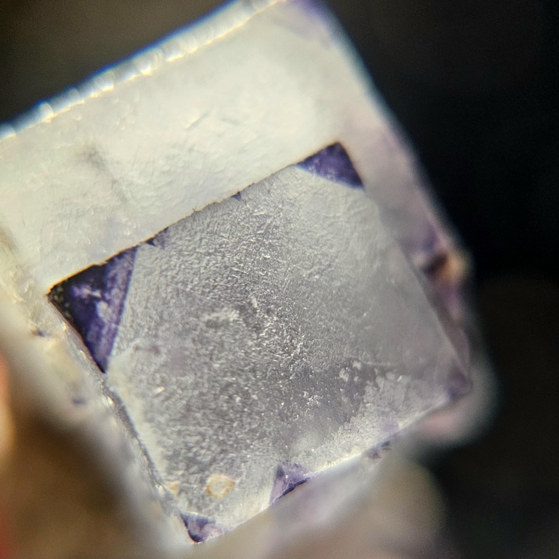 K&ouml;nnte beinhalten: Nahaufnahme eines durchscheinenden, kubischen Mineralexemplars mit mattiertem Aussehen. Der W&uuml;rfel hat eine helle, fast wei&szlig;e Farbe mit tiefvioletten Flecken an den Ecken. Die Oberfl&auml;che hat ein strukturiertes, kristallines Aussehen, was auf eine nat&uuml;rliche Bildung hindeutet.