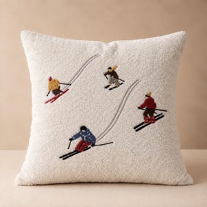 May include: A square, cream-coloured, textured throw cushion featuring an embroidered design of four skiers in motion. The skiers wear colourful winter gear, including red, blue, brown, and yellow jackets, and are depicted on skis with poles.