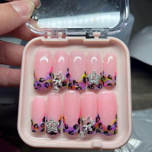 May include: A set of ten artificial nails in a pink case. The nails are pink with glitter and a leopard print design in purple, black, yellow, and orange. Each nail has a silver star charm.