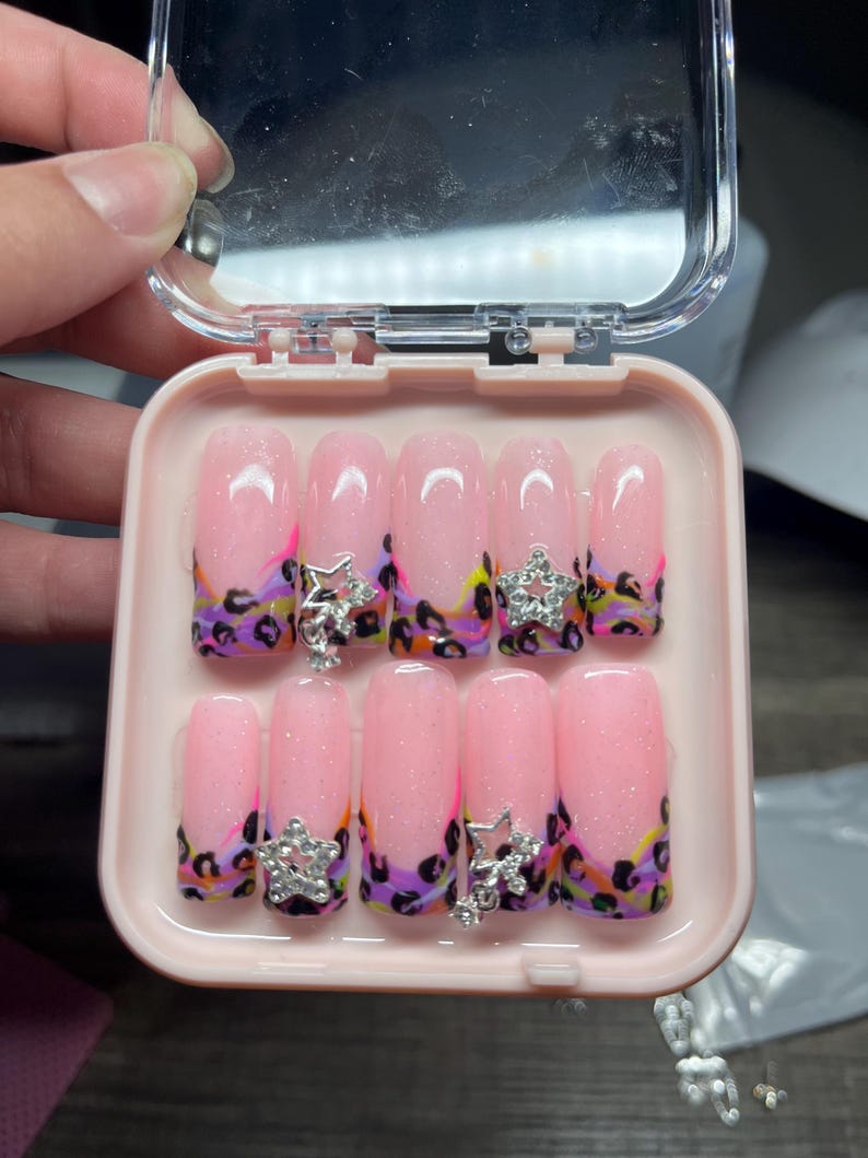 May include: A set of ten artificial nails in a pink case. The nails are pink with glitter and a leopard print design in purple, black, yellow, and orange. Each nail has a silver star charm.