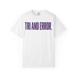 Könnte beinhalten: Ein weißes kurzärmeliges T-Shirt mit dem Text "TRI AND ERROR." in dunkelvioletten Großbuchstaben auf der Brust, wobei ein kleiner roter Punkt nach dem Wort "ERROR." platziert ist. Das Shirt hat einen Rundhalsausschnitt und eine Standardpassform.