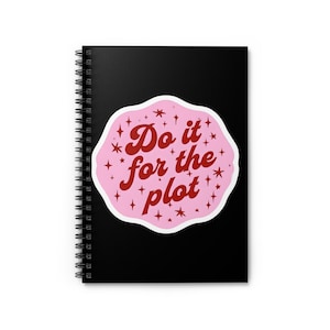 May include: Black spiral-bound notebook featuring a pink sticker with the text "Do it for the plot" in red script, surrounded by stars. The notebook's cover is a solid black colour.