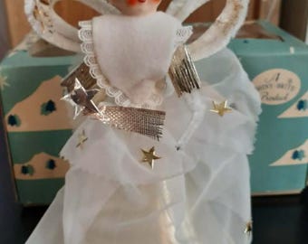 May include: A vintage angel tree topper featuring a painted face with rosy cheeks, wearing a white fabric gown with gold star accents. The figure has white wings with gold trim, a lace collar, and a sheer veil. A metallic star wand is held in hand.