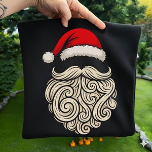 May include: Black square cushion featuring a Santa Claus design. The design includes a red Santa hat with white trim and a fluffy white pom-pom. Below the hat is a stylized white beard and mustache.