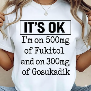 Może przedstawiać: Biała koszulka z napisem "IT'S OK" w czarnym prostokącie. Poniżej koszulka głosi "I'm on 500mg of Fukitol and on 300mg of Gosukadik" czcionką w kolorze czarnym. Koszulka jest trzymana przez osobę.