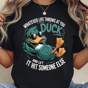 May include: Black t-shirt featuring a cartoon duck wearing sunglasses, with the text "Whatever life throws at you, duck and let it hit someone else." The duck is teal with an orange beak and feet. The text is in white and teal.