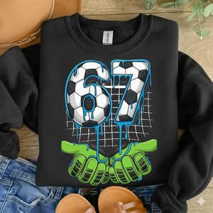 May include: Black sweatshirt with a soccer ball design featuring the number 67 in white and black, with blue dripping accents. The design includes green hands holding the number. A fun, sporty design.