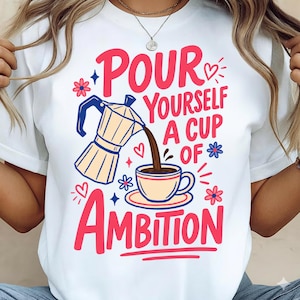 May include: A white t-shirt featuring a graphic print of a moka pot pouring coffee into a cup. The text reads "POUR YOURSELF A CUP OF AMBITION" in bold red letters. The design includes small blue and red flower icons, hearts, and star accents.