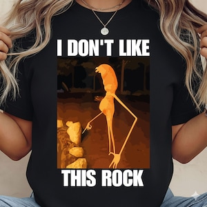 Puede incluir: Camiseta negra con el texto "I DON'T LIKE THIS ROCK." La camiseta presenta una figura de dibujos animados naranja que señala una roca. El fondo es marrón.