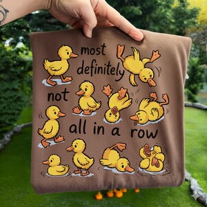 Op de afbeelding: Bruin vierkant kussen met gele cartoon eenden in verschillende poses, met de tekst "most definitely not all in a row". De eenden zijn in water en op het land.