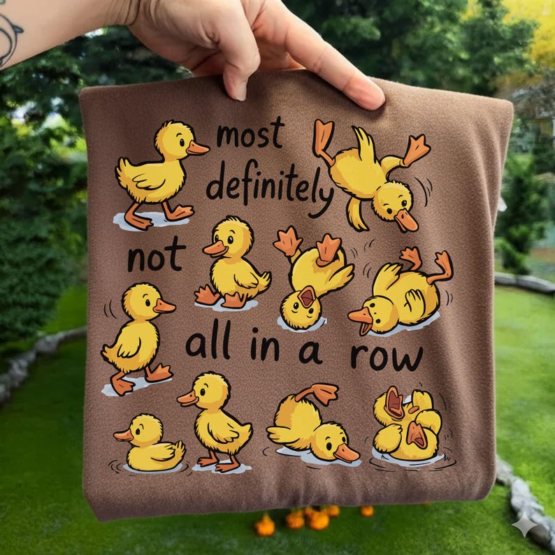 Op de afbeelding: Bruin vierkant kussen met gele cartoon eenden in verschillende poses, met de tekst "most definitely not all in a row". De eenden zijn in water en op het land.