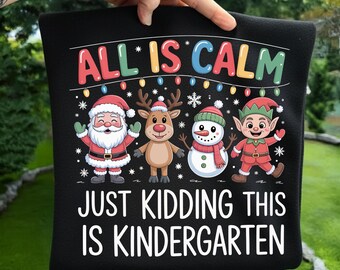 Könnte beinhalten: Schwarzes Kissen mit weihnachtlichem Design. Das Design zeigt den Weihnachtsmann, ein Rentier, einen Schneemann und einen Elf, mit dem Text "ALL IS CALM" und "JUST KIDDING THIS IS KINDERGARTEN".