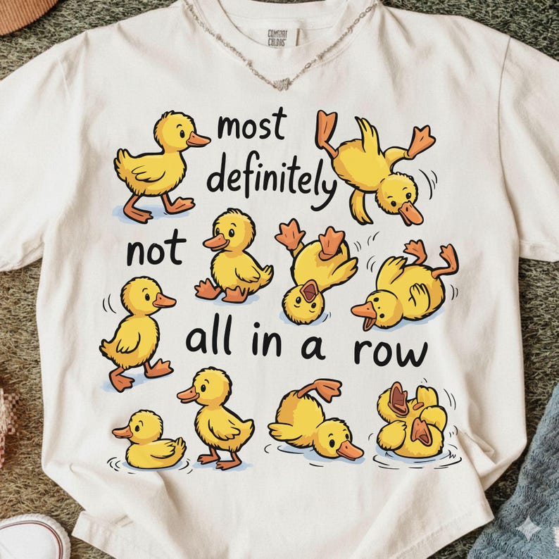 Op de afbeelding: Cr&egrave;mekleurig T-shirt met cartoongele eendjes in verschillende poses. De tekst op het shirt luidt "most definitely not all in a row". Het ontwerp is speels en grillig, perfect voor vrijetijdskleding.