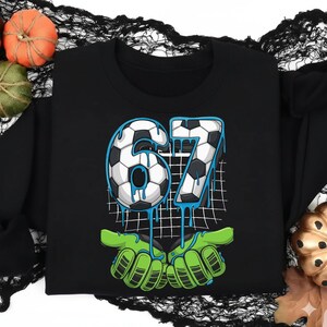 May include: Black sweatshirt featuring the number "67" in a soccer ball design with blue dripping accents. The number is held by green zombie-like hands. Decorative pumpkins and black lace are in the background.