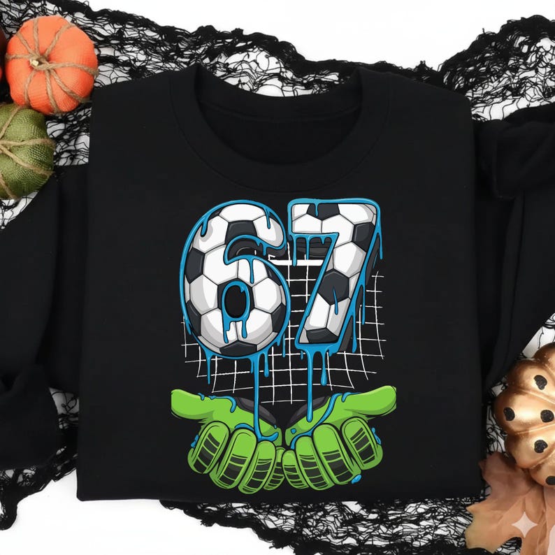 May include: Black sweatshirt featuring the number "67" in a soccer ball design with blue dripping accents. The number is held by green zombie-like hands. Decorative pumpkins and black lace are in the background.