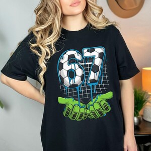 May include: Black t-shirt featuring a soccer-themed graphic. The design includes the number "67" in a soccer ball pattern with blue dripping accents, held by green goalie gloves. A grid pattern is in the background.