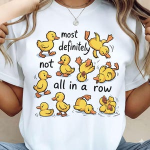 May include: White t-shirt featuring a playful design of yellow cartoon ducks in various poses. The text "most definitely not all in a row" is also included. The ducks are depicted in different positions, adding a whimsical touch to the shirt.