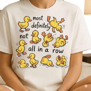 Op de afbeelding: Wit T-shirt met een speels ontwerp van gele cartoon eendjes in verschillende poses. De tekst "most definitely not all in a row" is boven en onder de eendjes gedrukt. Het shirt heeft korte mouwen en een ronde hals.