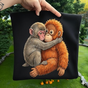 May include: Black square pillow featuring a heartwarming illustration of two monkeys embracing. One monkey is gray with a pink face, and the other is orange. The design is set against a solid black background.