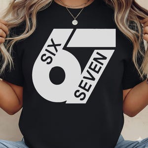 May include: Black t-shirt with a large white graphic design. The design features the word "SIX" in a curved font, and the word "SEVEN" in a slanted font, with the numbers 6 and 7 integrated into the design.