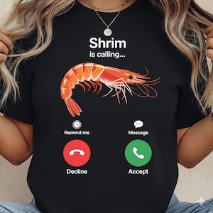 Op de afbeelding: Zwart T-shirt met een afbeelding van een garnaal en de tekst "Shrimp is calling...". Onder de garnaal staan de woorden "Remind me", "Decline", "Message" en "Accept" met bijbehorende iconen.
