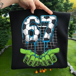 May include: Black square pillow with a soccer-themed design. The number "67" is depicted with a soccer ball pattern and blue dripping accents. Green goalie gloves are at the bottom.